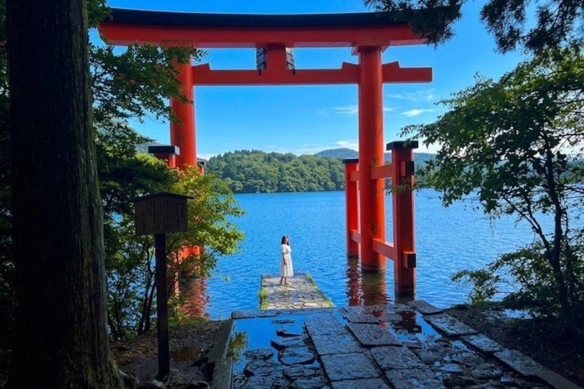 A red torii gate, a person standing on a stone platform, and a lake with trees in the background. 2