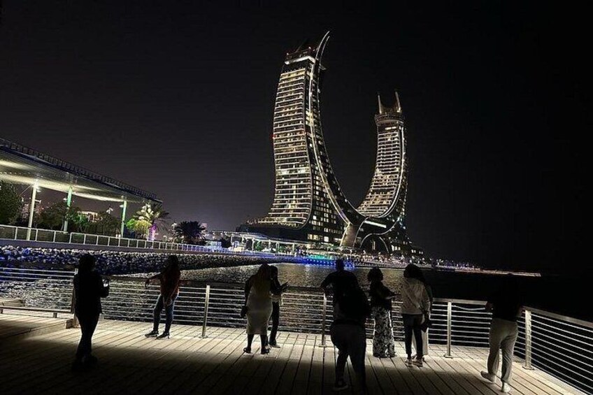 Vista nocturna de un puente grande e iluminado con dos torres, una pasarela de madera y gente de pie sobre ella. 4