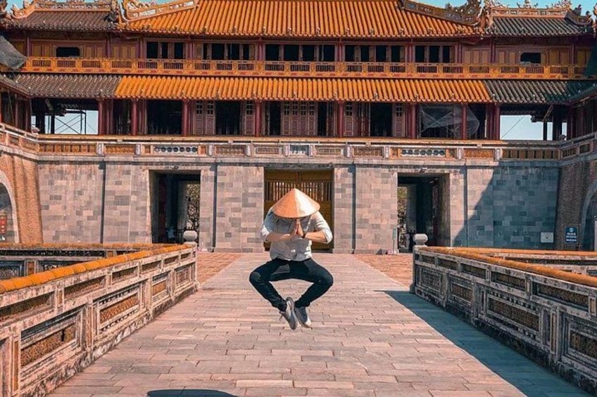 Una persona con cappello a forma di cono che esegue un salto di fronte a un edificio tradizionale con tetto in tegole. 1