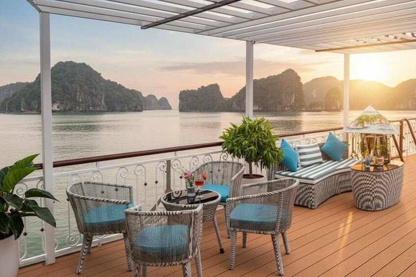 A wooden deck with wicker furniture, a table with drinks, and a view of a body of water with mountains in the background. 4