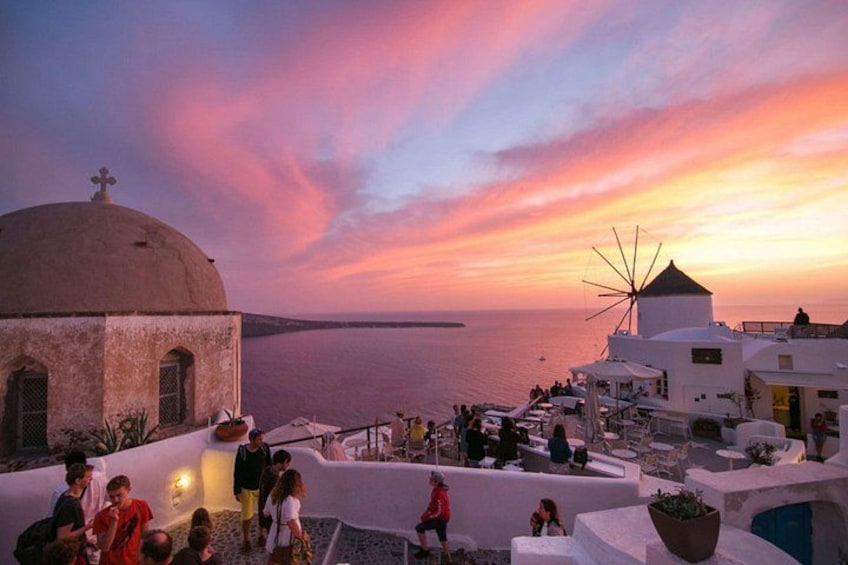 A sunset over a coastal village with white buildings, a windmill, and people gathered on the waterfront. 5