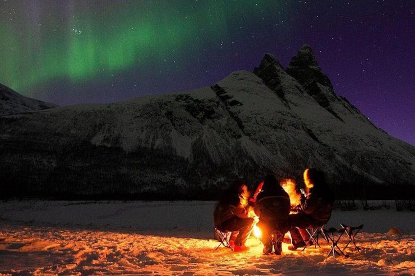 Eine Gruppe von Menschen sitzt an einem Lagerfeuer in verschneiter Landschaft mit Bergen im Hintergrund. 3