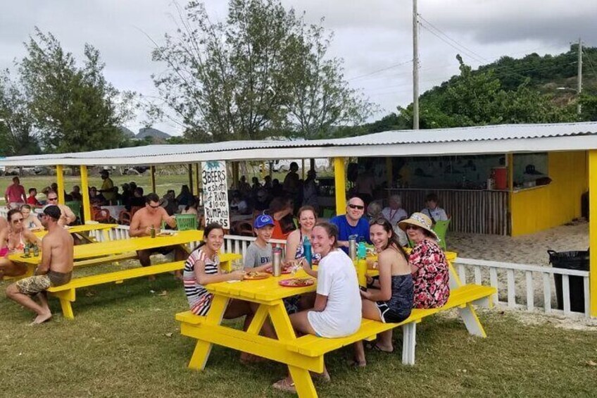 People sitting at yellow picnic tables under a covered area with a sign that reads "3 BEERS". 2