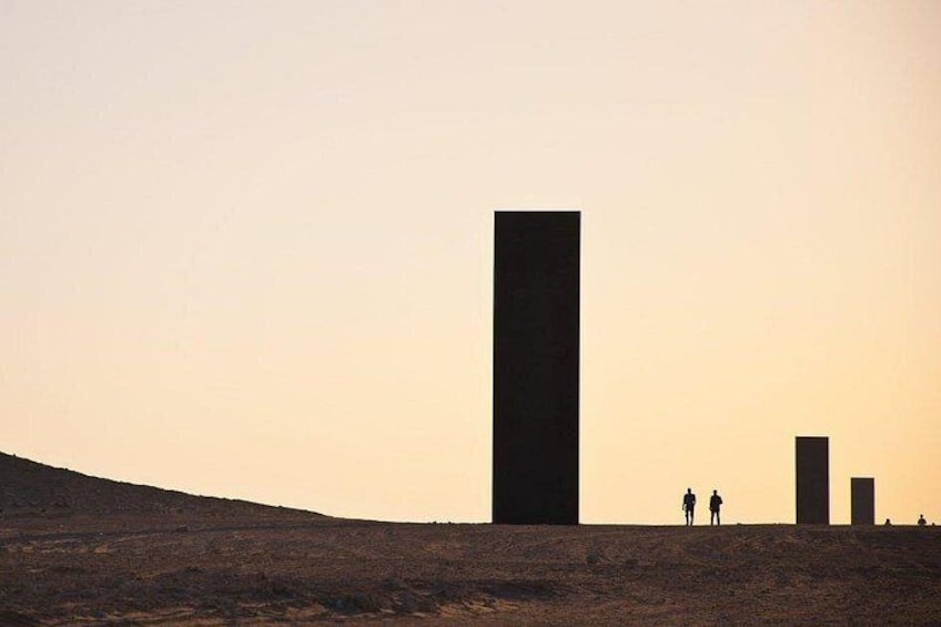 A desert landscape with three large vertical structures and two people walking. 5