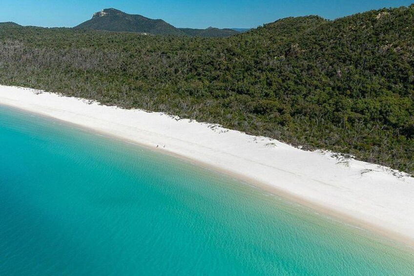 Una playa con agua turquesa cristalina, arena blanca y una colina boscosa al fondo. 2
