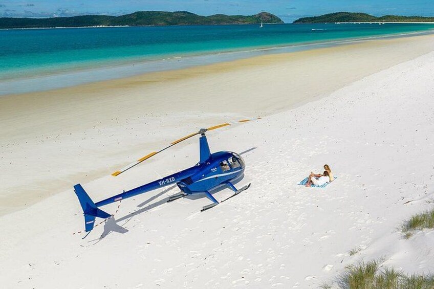 Un helicóptero azul sobre una playa arenosa con una persona sentada cerca, sobre una toalla. 4