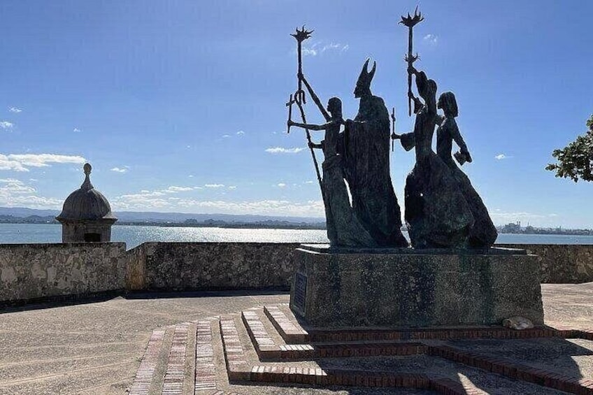 Une statue représentant trois personnages, placée sur un socle, avec un plan d’eau et un dôme en arrière-plan. 4