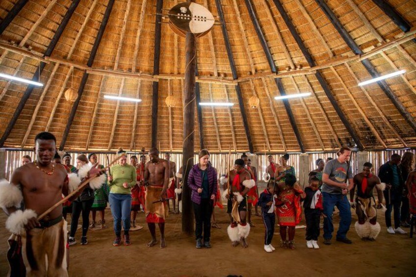 A group of people in a thatched-roof structure, some holding traditional instruments. 4