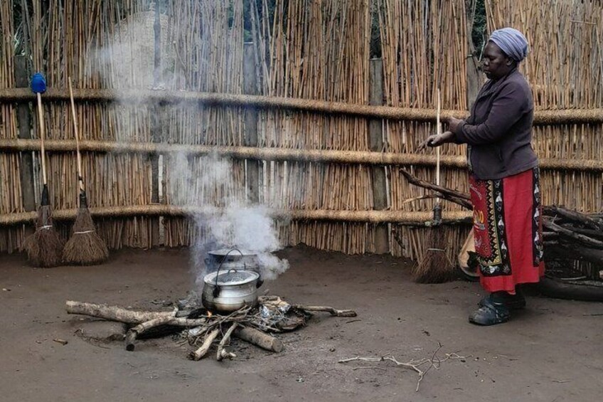 Iemand kookt boven een open vuur met een pan, omringd door een rieten muur en traditionele bezems. 5