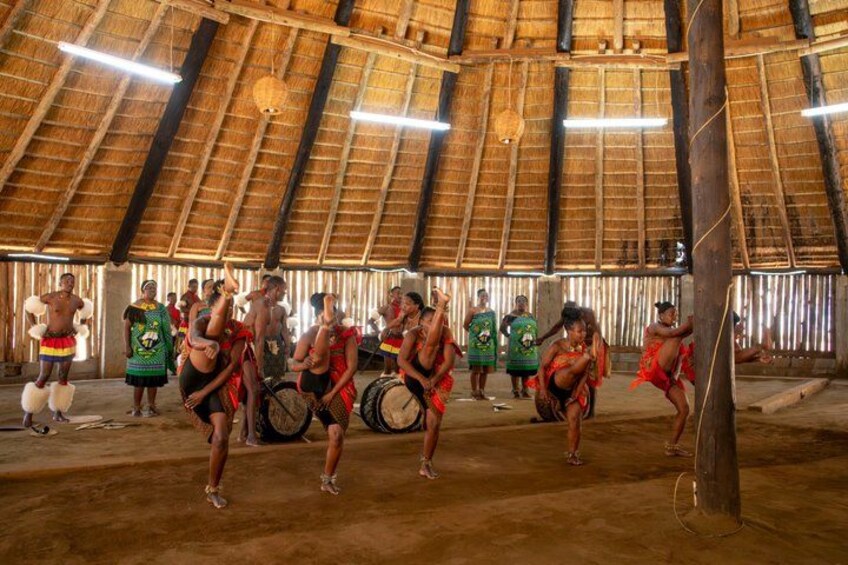 A group of people in traditional attire performing a dance in a thatched-roof hut. 1