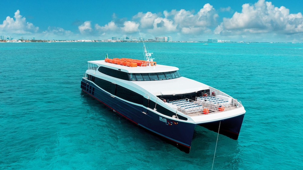 Een groot cruiseschip genaamd XEL-HA vaart over turquoise water, met een stadsachtergrond op de achtergrond. 1