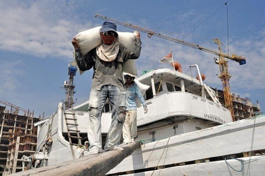 Un trabajador en un barco con un saco blanco grande, una grúa y construcción al fondo. 3