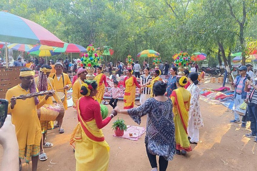 A group of people in traditional attire, possibly at a cultural event, with trees and colorful umbrellas in the background. 2