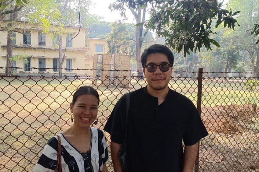 Two people standing in front of a chain-link fence with a building and trees in the background. 5