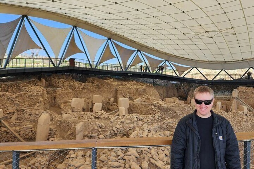 A person standing in front of an archaeological site with exposed ruins and a modern shelter overhead. 4
