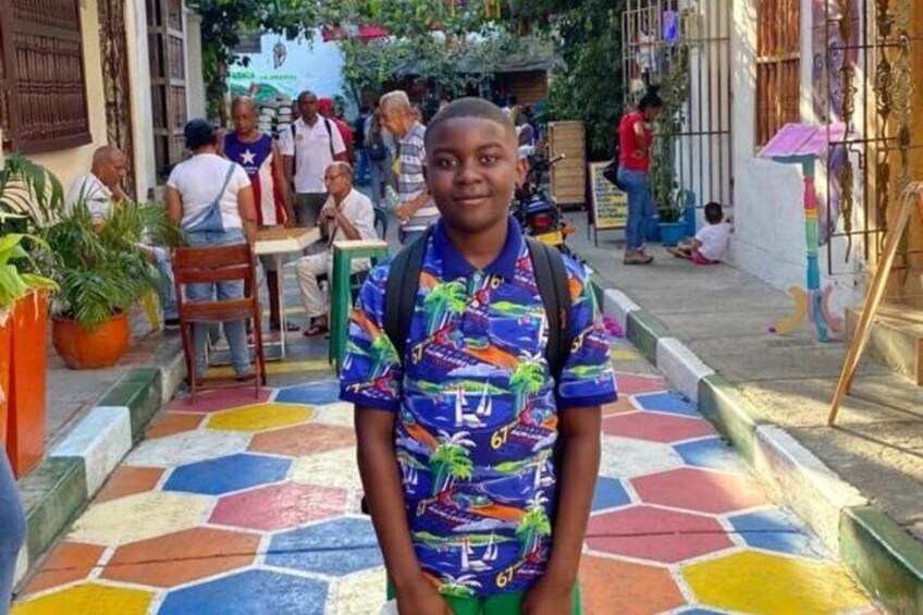Una persona de pie sobre un pavimento de baldosas de colores, con una mochila, rodeada de gente y edificios. 5