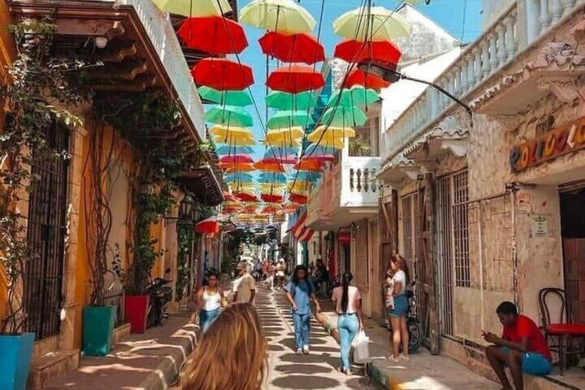 Una calle estrecha con sombrillas de colores colgando sobre ella, y gente caminando y sentada. 2