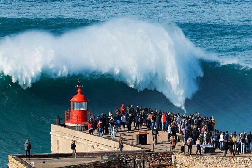 A large wave crashing over a lighthouse, with a crowd of people watching from the shore. 1