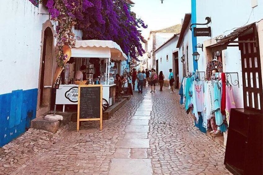 A cobblestone street with a food cart, people walking, and colorful clothing on display. 2