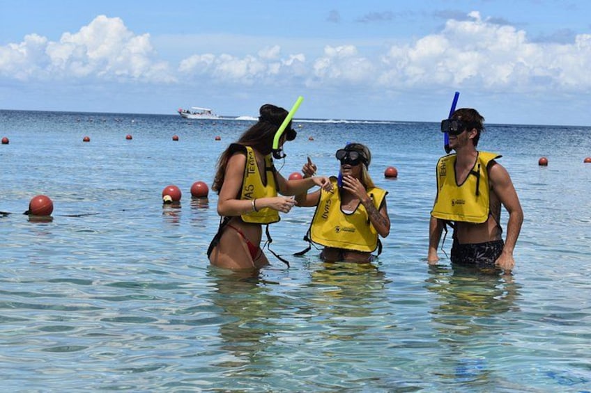 Tres personas con chalecos salvavidas amarillos y snorkels en aguas poco profundas, con boyas y un bote al fondo. 4