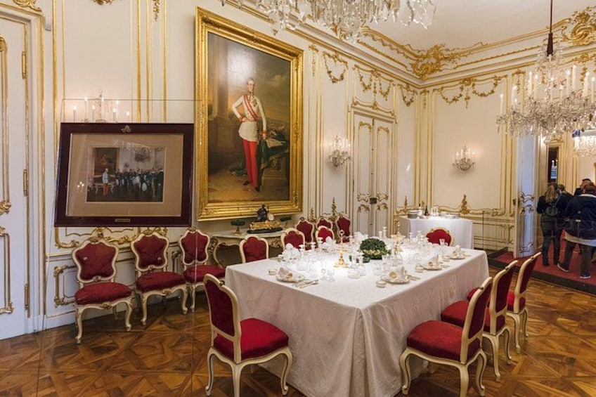 A formal dining room with a large table set for a meal, red upholstered chairs, ornate gold detailing, and a large portrait on the wall. 4