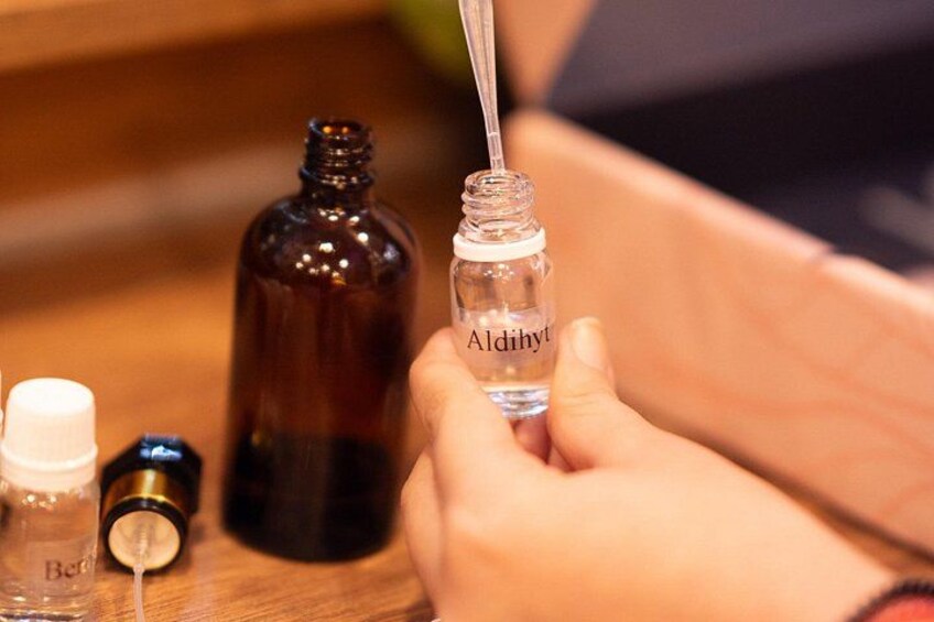 A hand holding a small bottle labeled 'Aldihyt' with a dropper, a brown glass bottle, and a white cap bottle on a wooden surface. 1