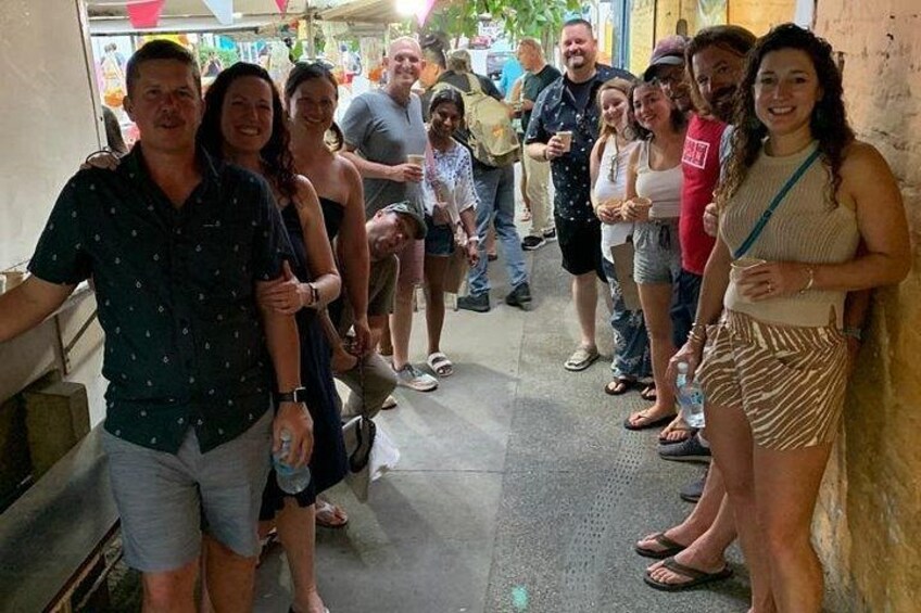 Un gruppo di persone in piedi in uno spazio esterno stretto, con pavimento in cemento, alcune con bevande in mano. 1