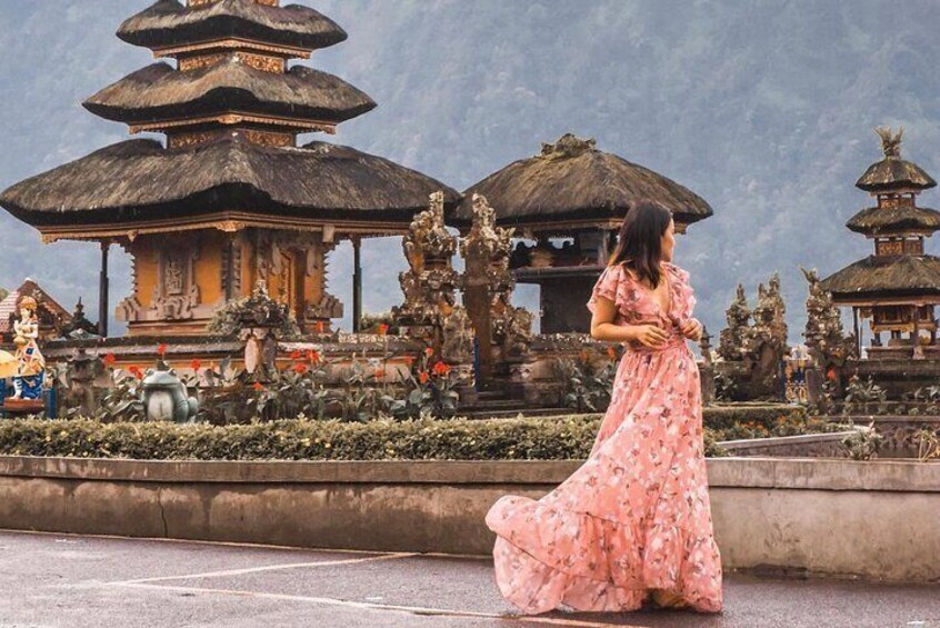 A woman in a pink floral dress standing in front of a traditional Balinese temple with tiered roofs and intricate statues. 4
