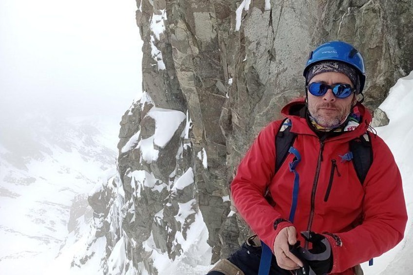 A person in a red jacket and blue helmet standing on a snowy mountain with rocky cliffs. 4