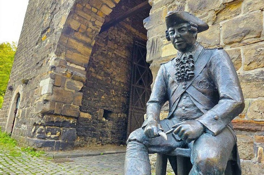 A statue of a historical figure seated on a bench, with a stone building in the background. 4