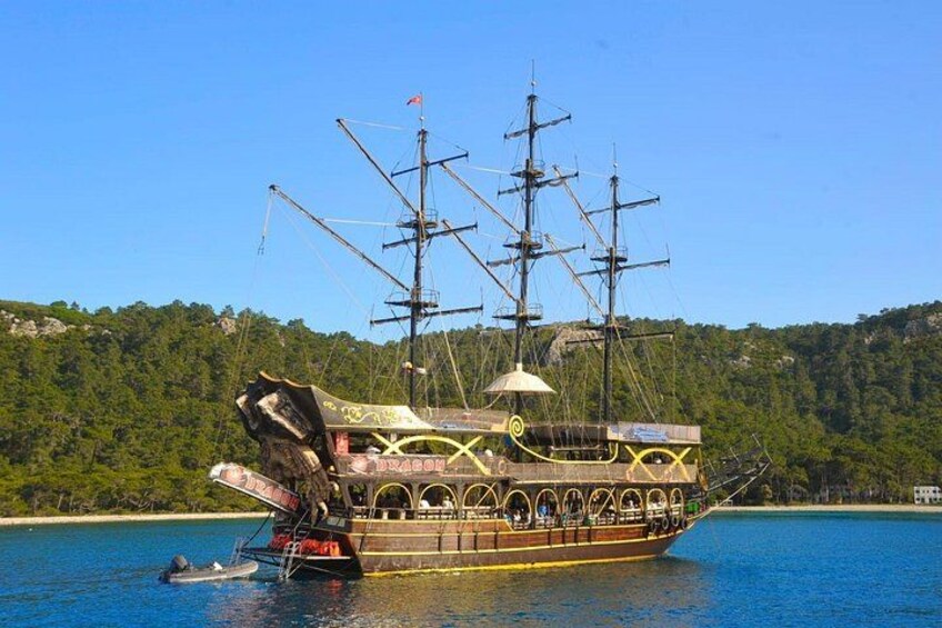 A large, ornate ship with multiple masts and a cabin, docked in a body of water with a forested shoreline in the background. 4