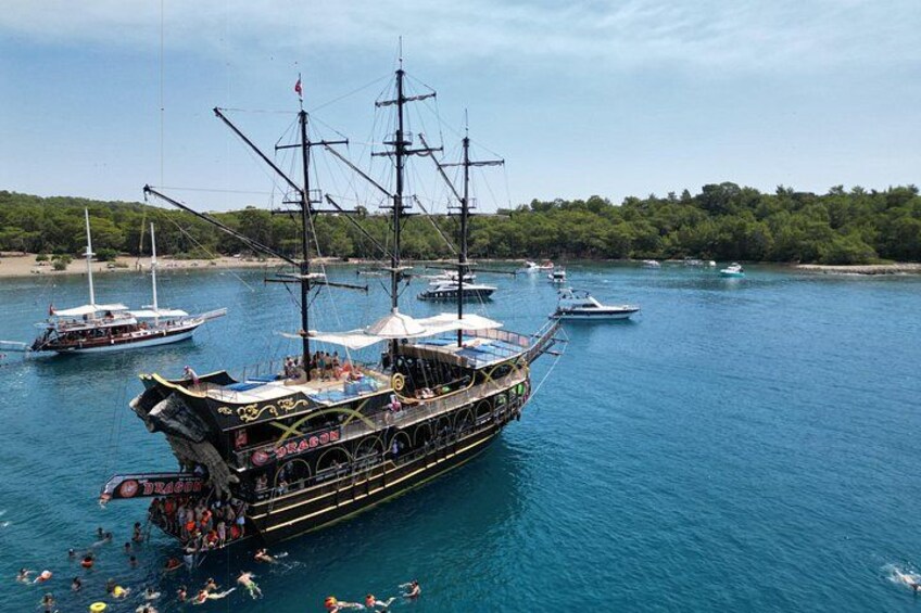 A large ship with multiple masts and passengers, docked in a harbor with other boats and a forested shoreline in the background. 2