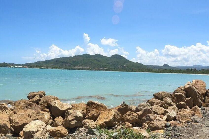 A rocky shoreline with clear turquoise water, distant green hills, and a clear sky. 3