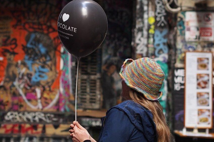 Una persona tiene un palloncino nero con la scritta 'I ❤️ CHOCOLATE', in piedi di fronte a un muro pieno di graffiti. 5