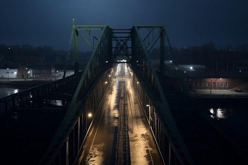 Een nachtzicht van een groene metalen brug met verlichting langs het voetpad, die naar een waterlichaam leidt. 2