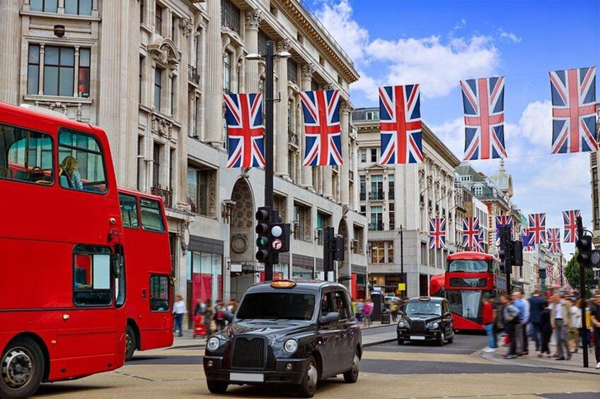 A red double-decker bus, a black car, and a red car on a city street with British flags. 4