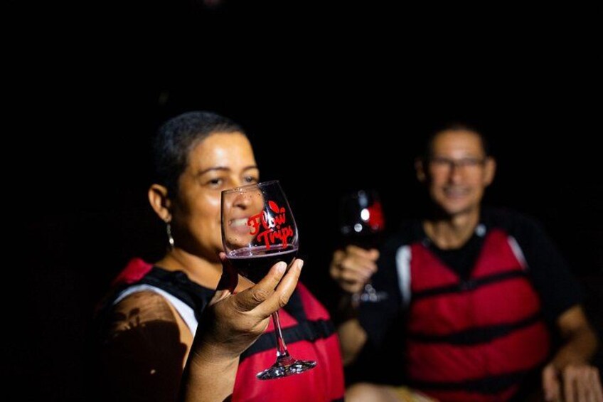 A person holding a glass with a red liquid, wearing a red life jacket, and another person in the background also wearing a life jacket. 4