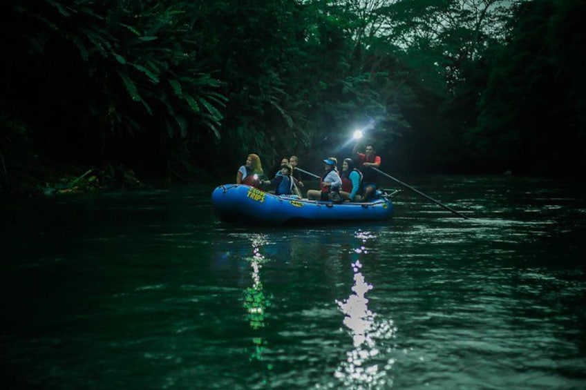 A group of people in a blue inflatable raft on a river surrounded by dense greenery. 1