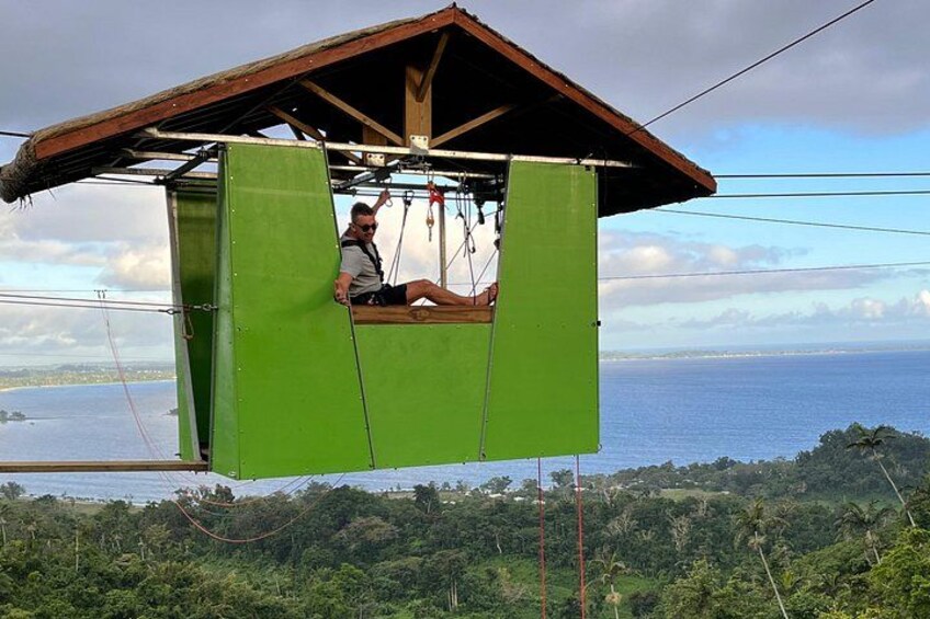 Une personne est assise dans un siège suspendu vert, sous un toit en bois, d’où elle a une vue imprenable sur un paysage côtier, avec une végétation luxuriante et l’océan. 2