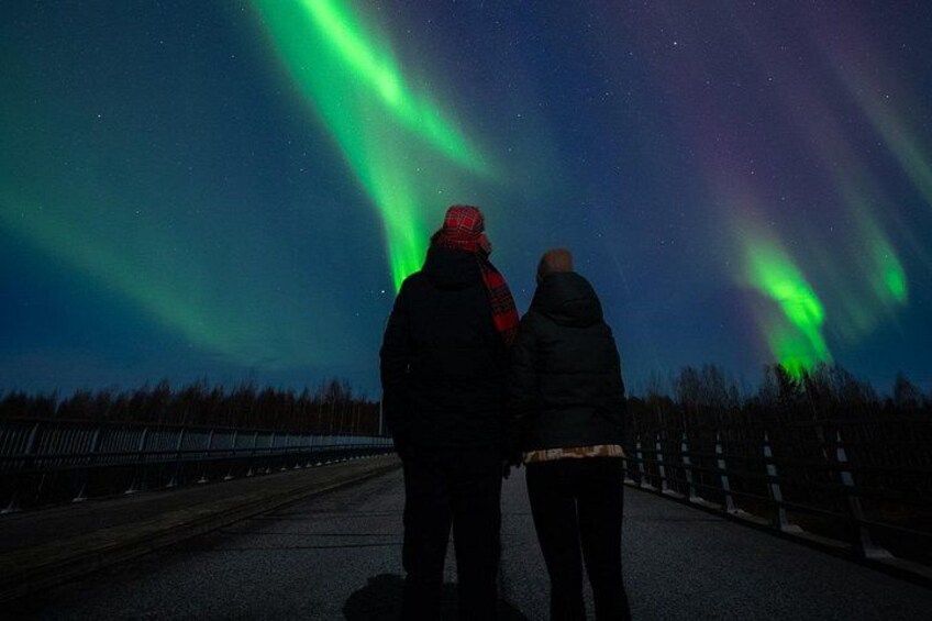 Dos personas de pie en una carretera, observando el cielo nocturno con auroras boreales. 4