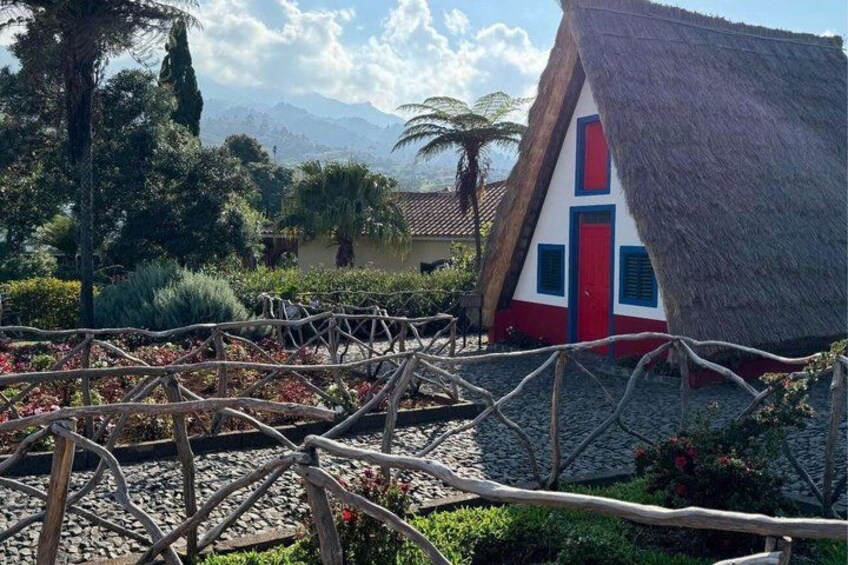 Une maison à toit de chaume, avec une porte rouge et des encadrements de fenêtres bleus, entourée d’un jardin délimité par une clôture en bois. 1