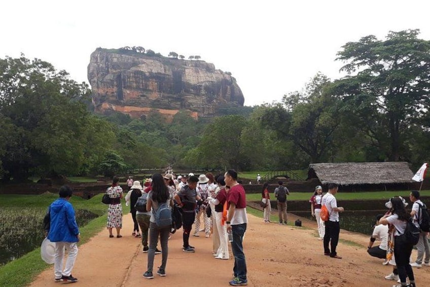 A group of people standing on a dirt path, a large rock formation in the background, and a thatched-roof structure to the right. 3