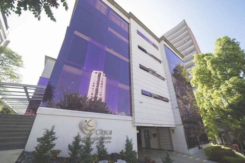Un edificio moderno con ventanas de vidrio reflectante, un letrero que dice 'Clínica' y una zona ajardinada con árboles. 3
