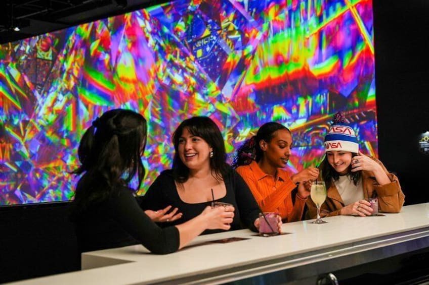 Four people sitting at a table with drinks, a colorful abstract background, and a NASA logo on a headband. 2