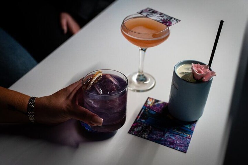 A person holding a purple drink with a lemon slice, a cocktail in a glass, and a blue drink with a rose garnish on a coaster. 5