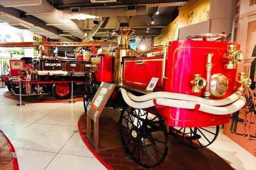 Un camión de bomberos rojo con detalles dorados y un carruaje antiguo en un museo. 3