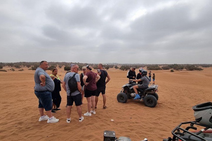 A group of people standing on a sandy terrain, with a quad bike in the foreground. 4