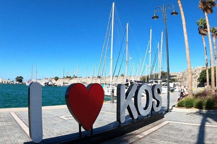 Un port de plaisance avec des bateaux, une pancarte sur laquelle on peut lire « J’aime KOS », et un ciel bleu azur. 2