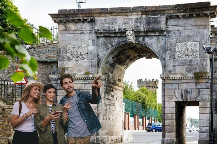 A stone archway with carvings, a castle in the background, and a blue car parked nearby. 1