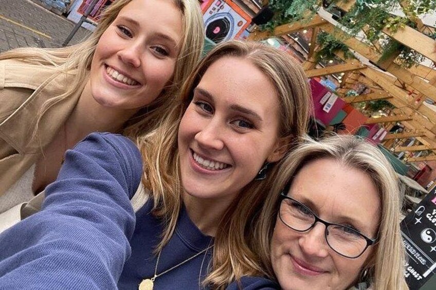 Three people taking a selfie in an outdoor setting with a wooden structure and colorful signs in the background. 2
